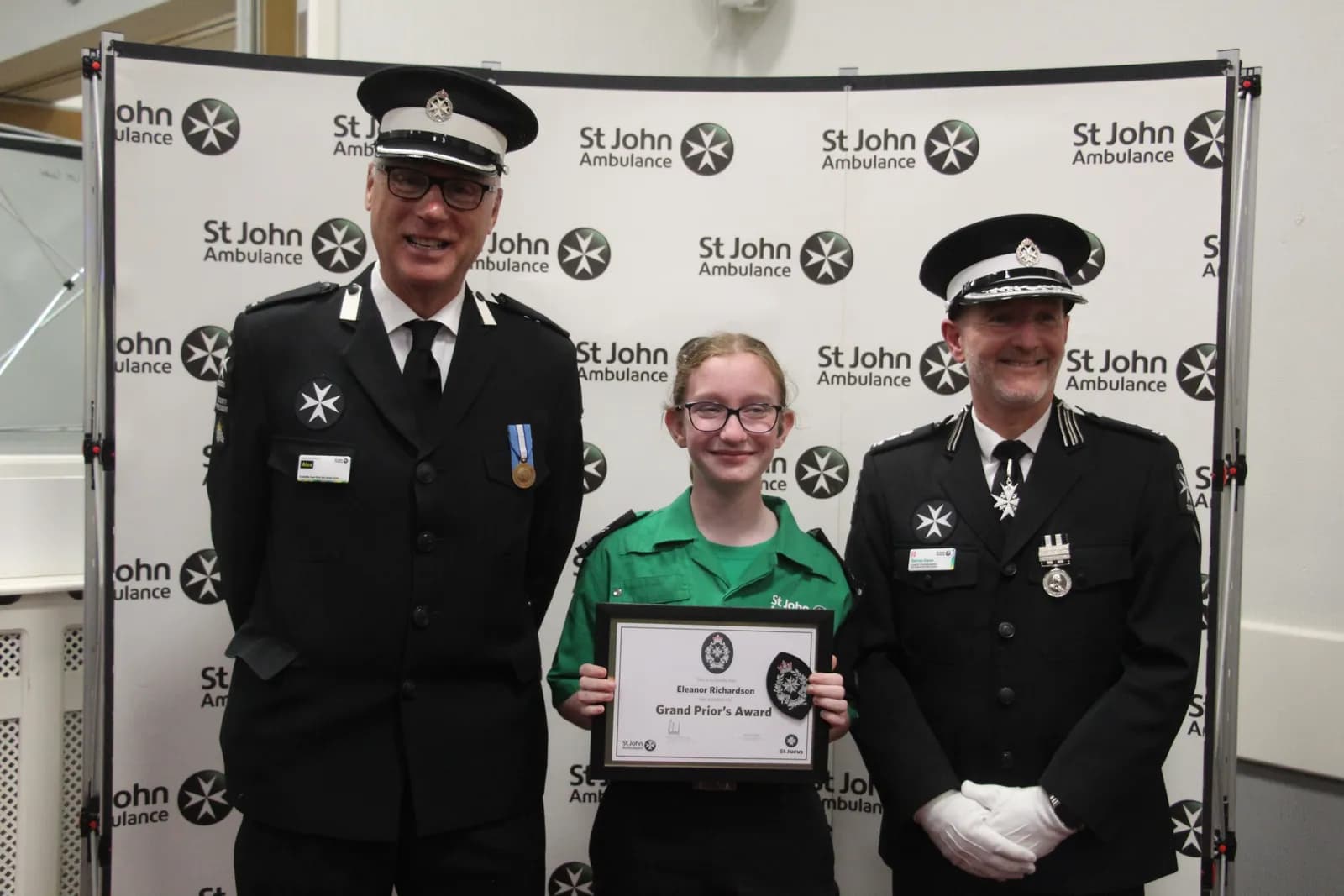 A young girl in a green uniform and glasses smiles while holding a framed certificate, flanked by two men in black uniforms with medals against a white backdrop featuring the St John Ambulance logo.